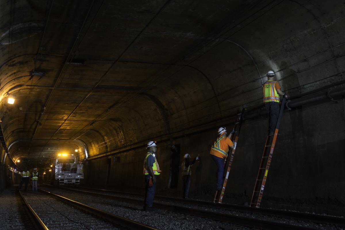 Workers in safety gear install cable brackets on subway tunnel walls under bright work lights.