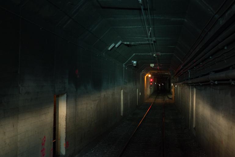 Dimly lit underground tunnel with railroad tracks and concrete walls, leading toward a distant lit doorway.