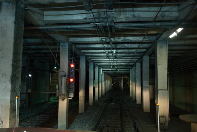 Dimly lit underground transit tunnel with concrete pillars, railway tracks, and red signal lights.