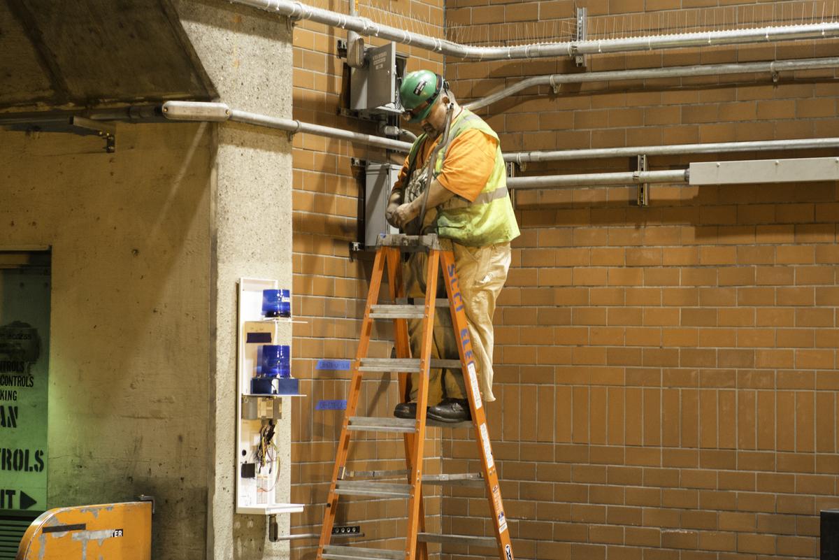Worker on ladder installing communication cable and brackets on brick subway station wall.