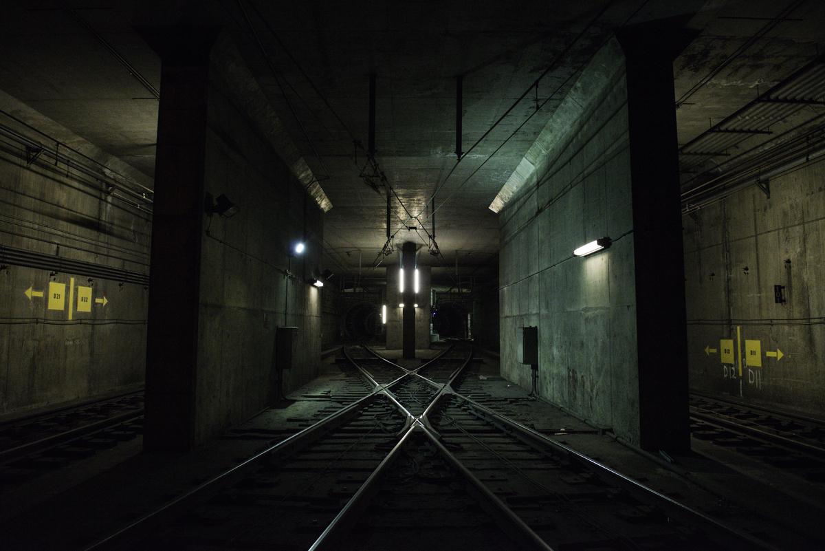 Underground train tunnel with crossing tracks and concrete walls lit by overhead lights.