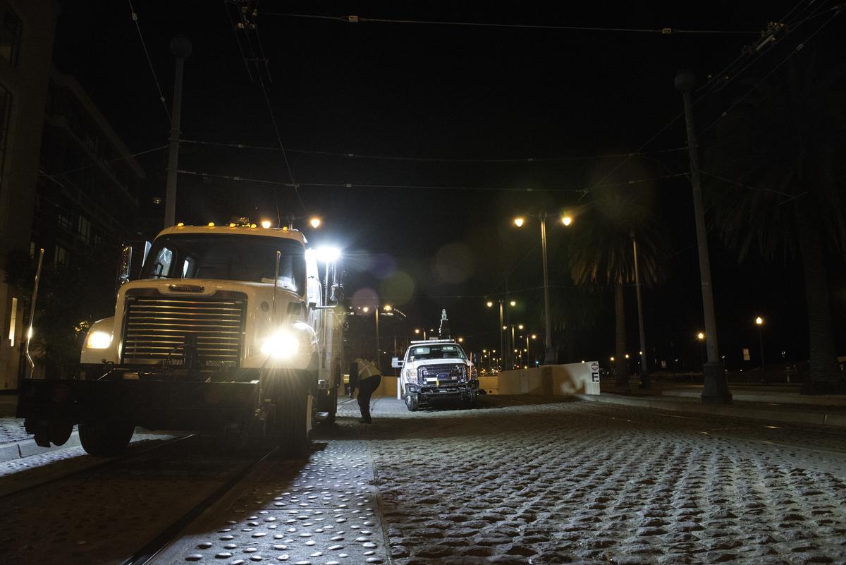 Construction trucks exiting Embarcadero Station tunnel at night on brick pavement with street lights.