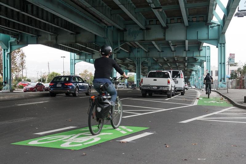 Cyclist riding in green-painted protected bike lane under elevated freeway with parked cars and traffic nearby.