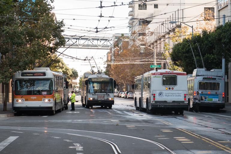 Multiple Muni buses, including vintage and modern models, pass each other on a street with overhead trolley wires.