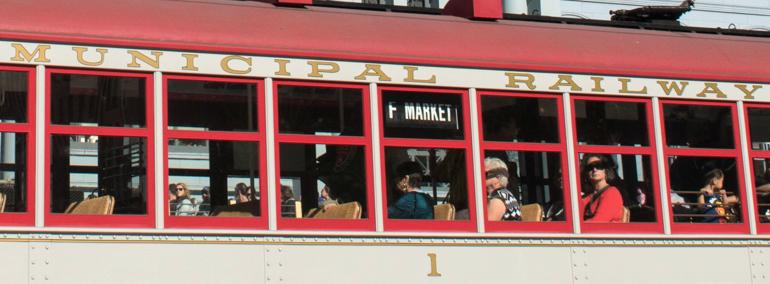 Municipal Railway streetcar with passengers visible through red-framed windows, gold lettering on cream-colored side panel.