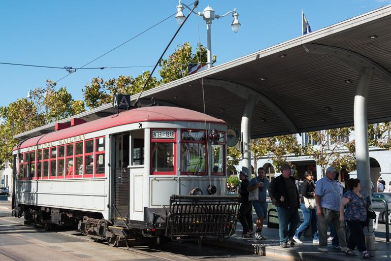 Red and white heritage streetcar with passengers boarding at covered transit station under clear sky.