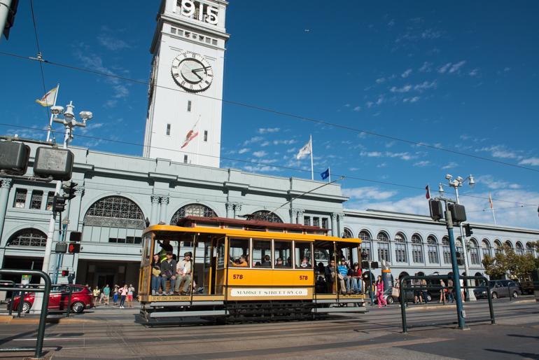 Vintage yellow streetcar 578 passes Ferry Building with clock tower under blue sky.