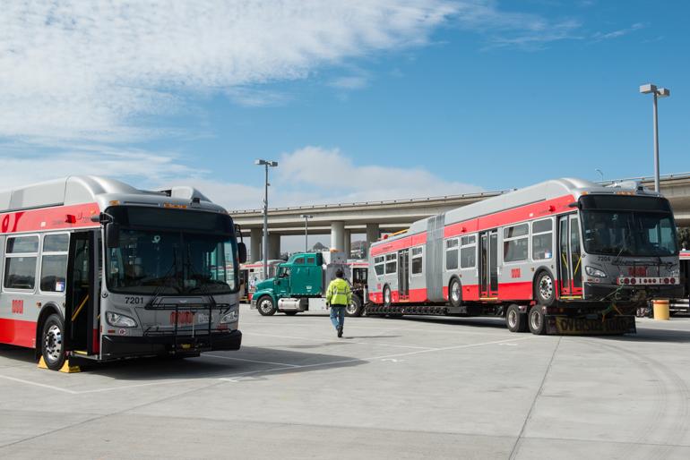Red and white articulated trolley coaches being delivered at Islais Creek Yard with tow truck and worker.