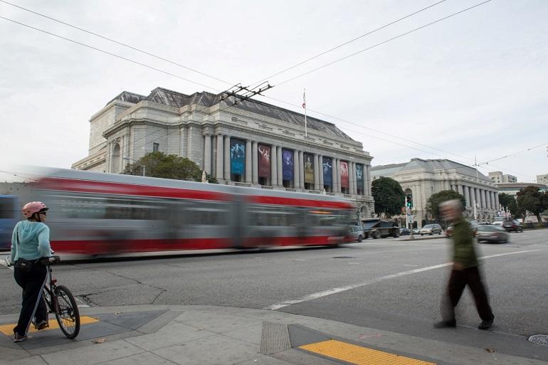 Red Muni bus on Van Ness Avenue with City Hall and pedestrians visible.