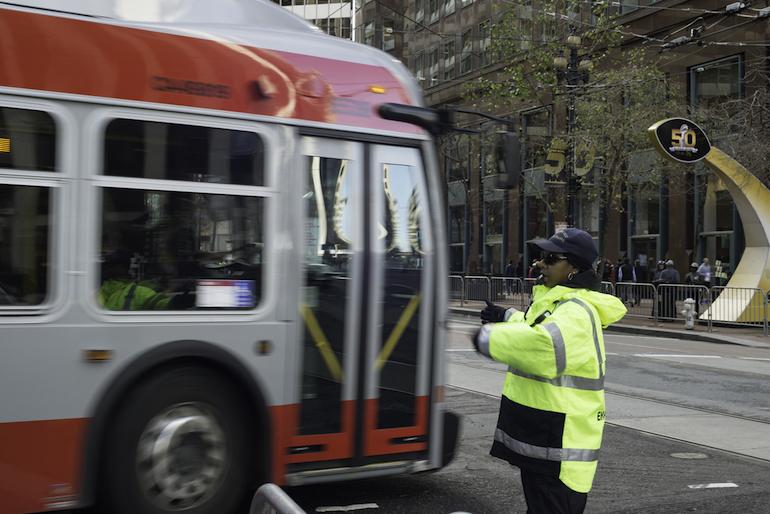 Traffic control officer in yellow safety vest directing red and gray Muni bus on Market Street.