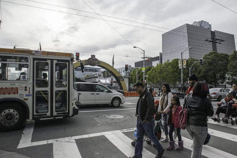 Pedestrians crossing Howard Street at a Muni bus stop with downtown San Francisco buildings in background.