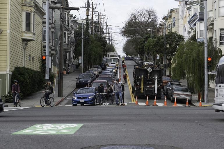 Street intersection with parked cars, construction equipment, traffic cones, and cyclists on a newly marked bike lane.