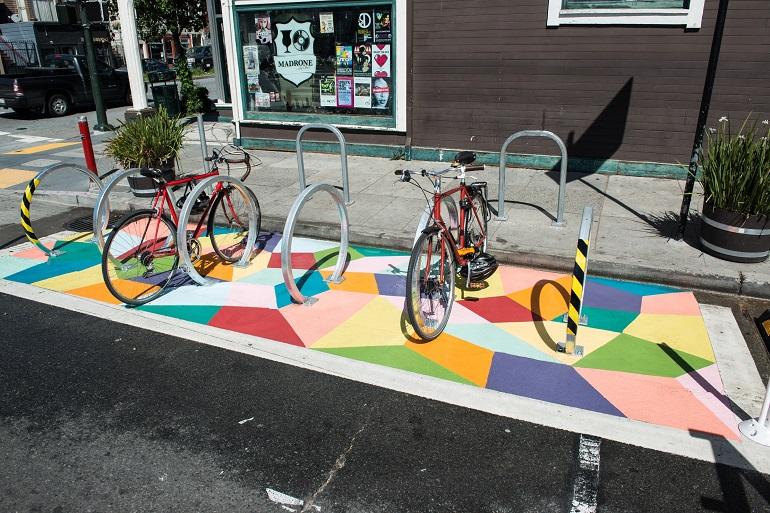 Six bicycles parked in racks on colorful geometric mural pavement in urban street setting.