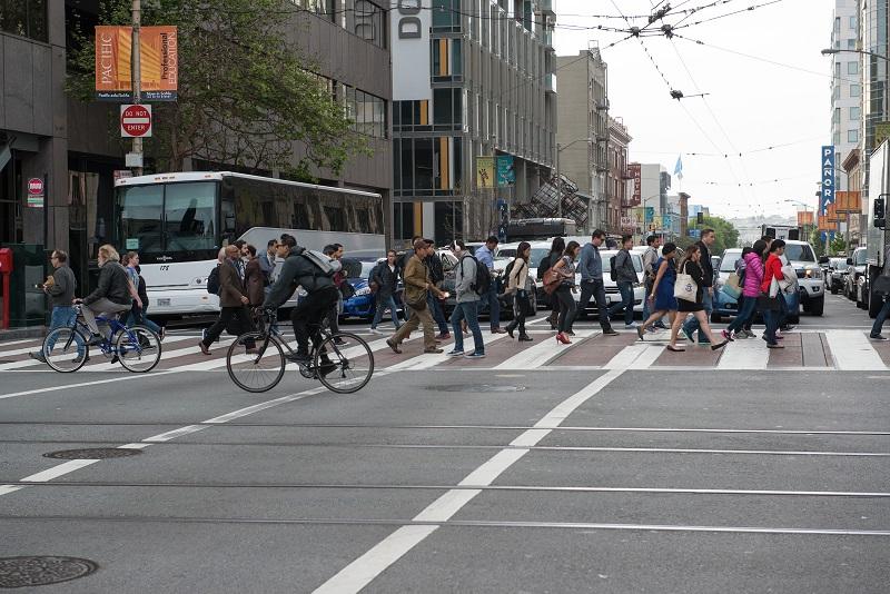 Commuter shuttle stopped at downtown intersection with pedestrians and cyclists crossing street.