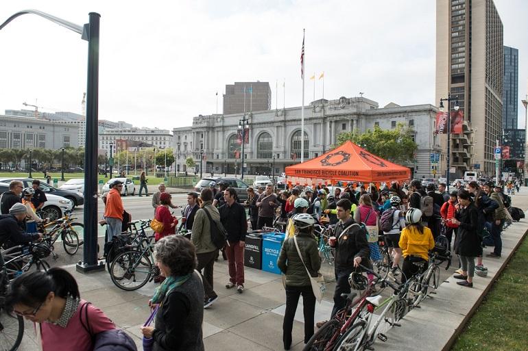 Crowd of cyclists and pedestrians gathered on sidewalk in front of City Hall with orange tent.