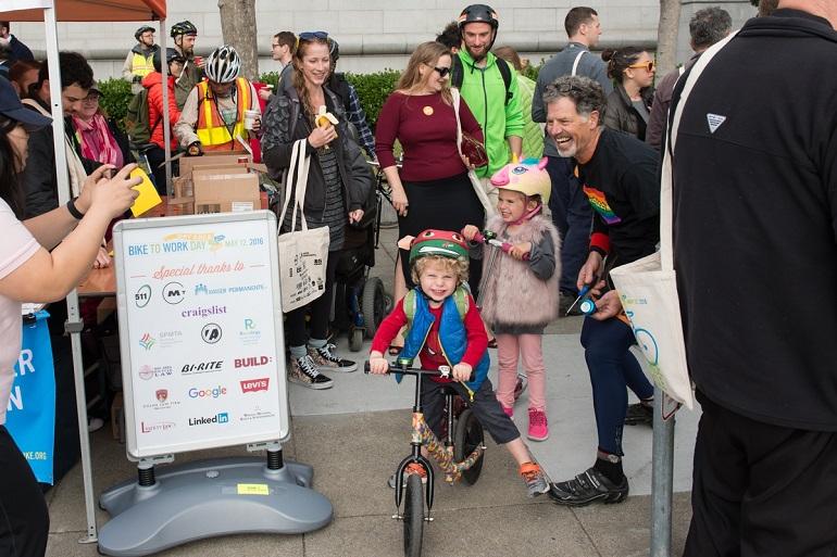 Young children with bike and scooter at Bike to Work Day event near City Hall with crowd of participants.