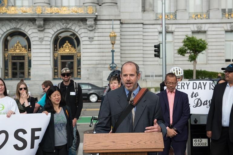 Man speaking at podium in front of City Hall with cyclists and officials behind him holding bike advocacy signs.