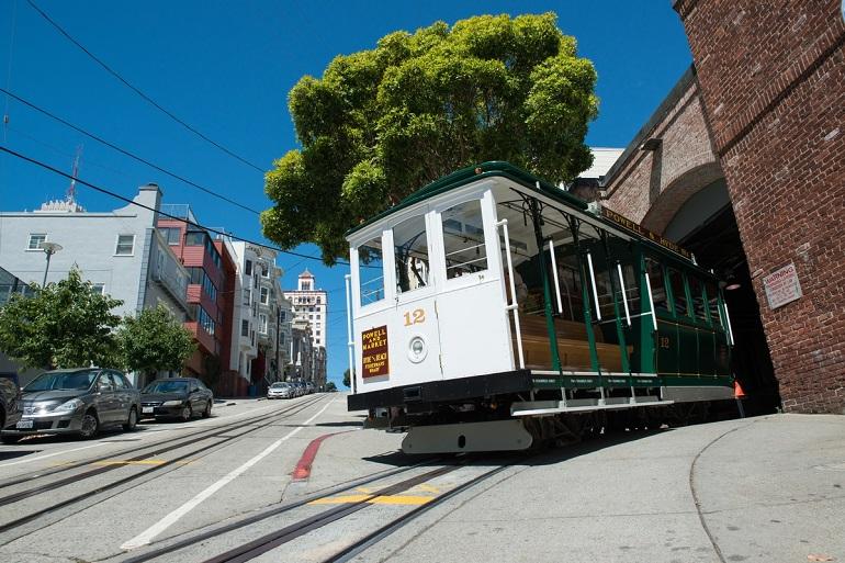 Cable Car 12 exits the red brick Cable Car Barn under clear blue sky on San Francisco street.
