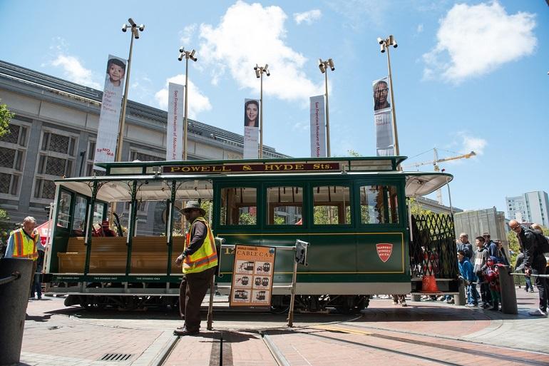 Green cable car numbered 12 on Powell & Hyde turntable with operators and pedestrians nearby.