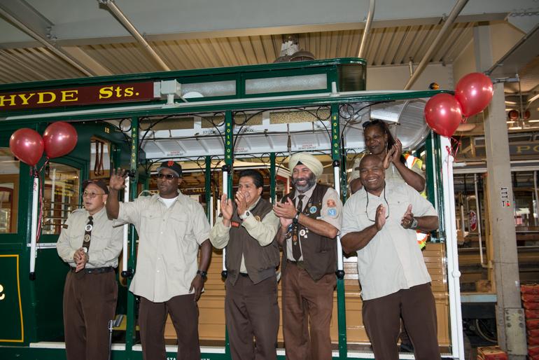 Seven people in uniform stand on a cable car with red balloons, clapping and smiling during a bell ringing contest announcement.