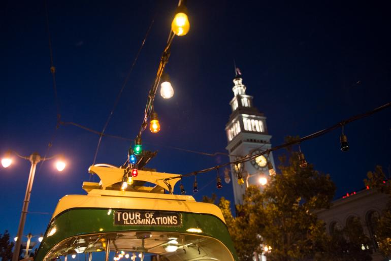 Streetcar 233 passes Ferry Building clocktower at night with illuminated lights and Tour of Illuminations sign visible.