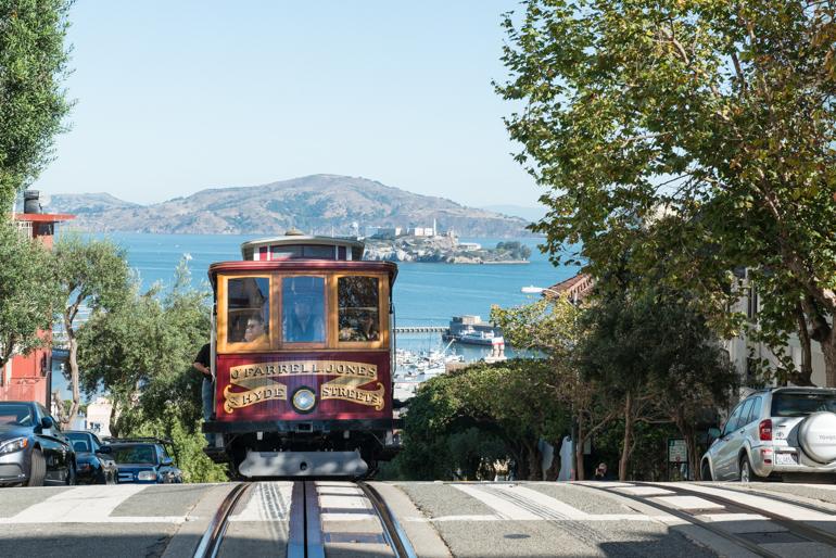 Red and gold cable car climbing steep hill with San Francisco Bay and Alcatraz Island visible in background.