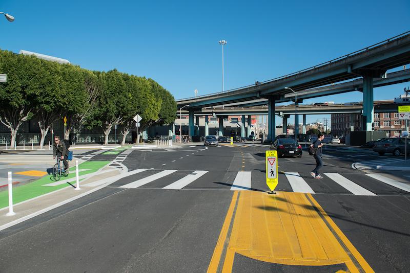 Tree-lined intersection with pedestrians crossing, bright yellow and green street markings, and highway overpass above.