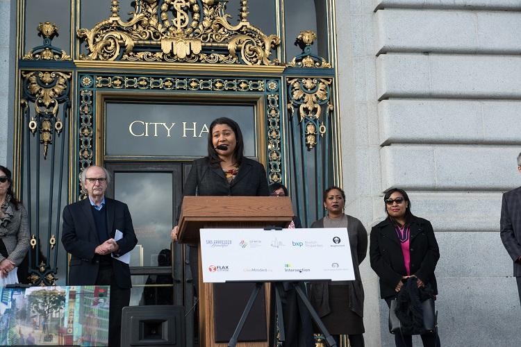 Woman speaking at podium in front of City Hall entrance during transit art program launch event.