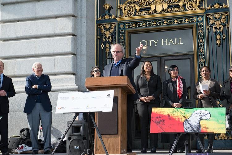Man speaking at podium outside City Hall with officials and colorful transit art display.