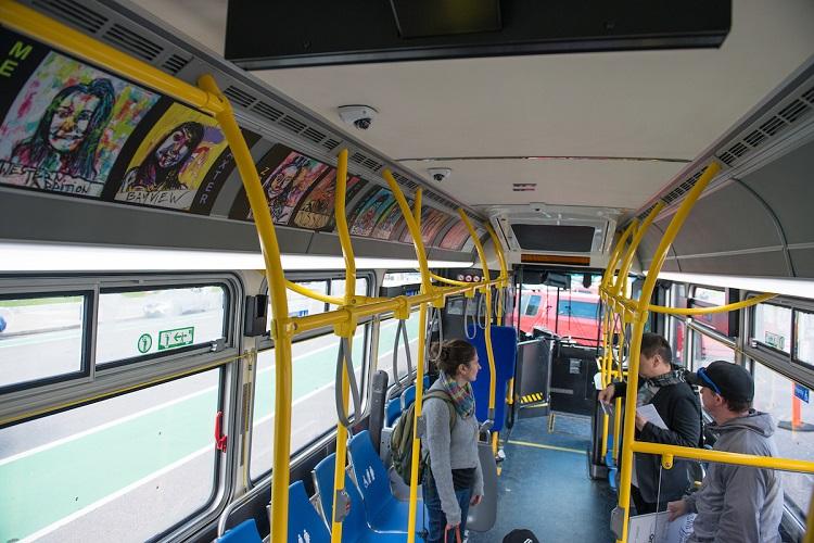 Interior of Muni bus with yellow handrails, blue seats, and colorful artwork displayed on upper panels above windows.