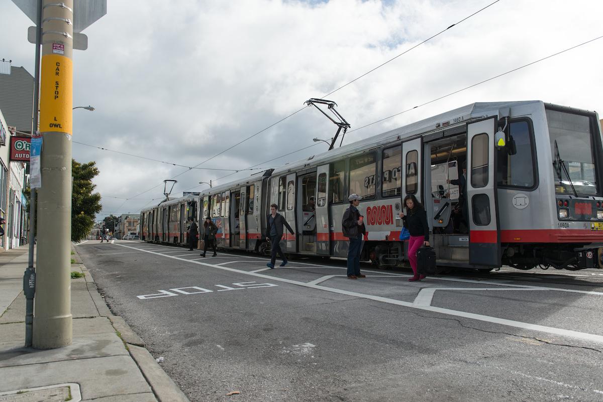 L Taraval light rail train stopped at station with passengers boarding and alighting near striped clear zone.