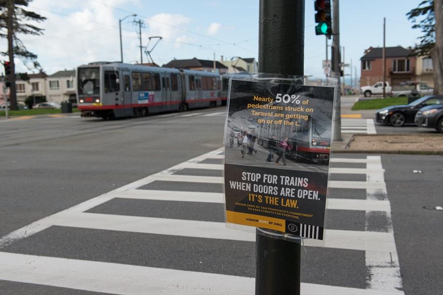 Safety campaign sign on pole at Taraval Street crossing with L-train visible in background.