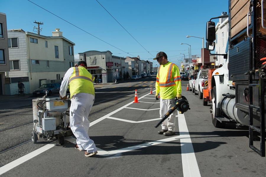 SFMTA workers in safety vests paint white street markings on Taraval Street.