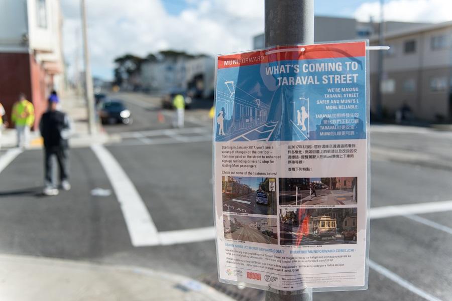 A sign posted on a utility pole announcing improvements coming to Taraval Street, with construction workers visible in the background.