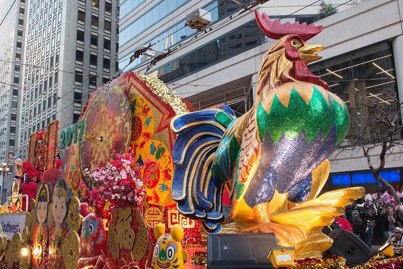 Colorful parade float featuring a tall glittery rooster with ornate decorative elements in downtown urban setting.