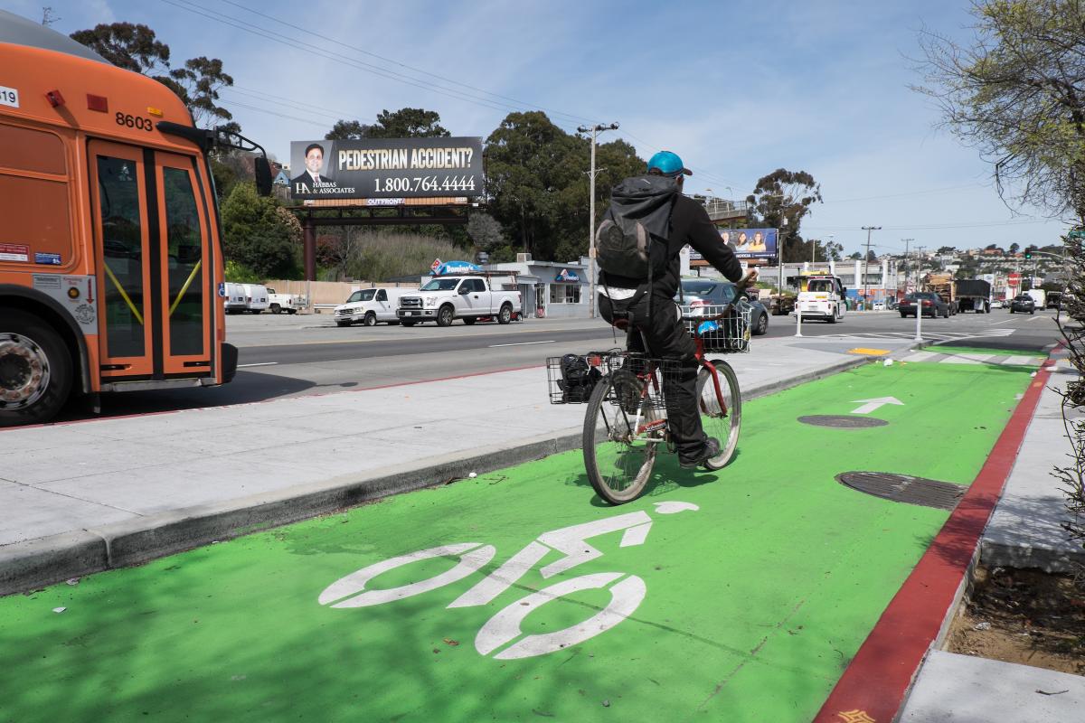 Cyclist riding on green protected bike lane next to orange transit bus on busy street.