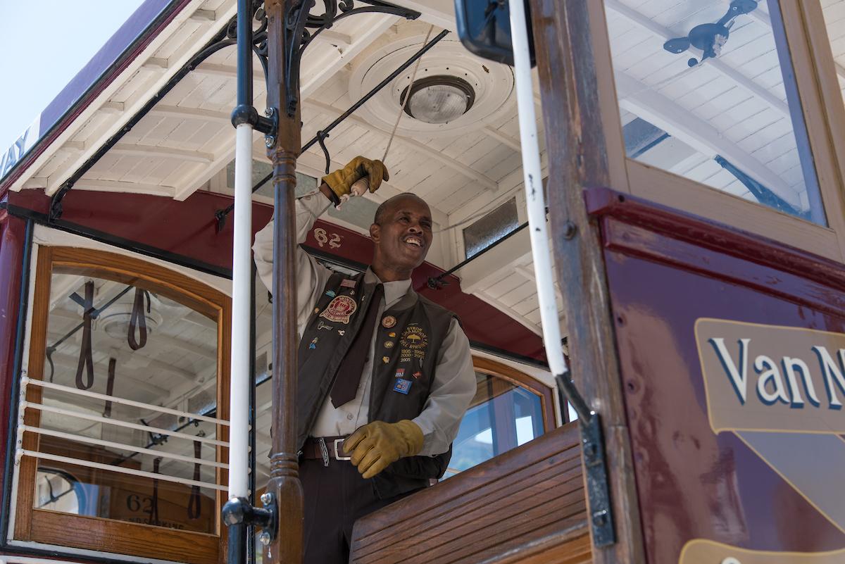 Muni cable car operator rings bell while standing in operator's cabin of vintage cable car.