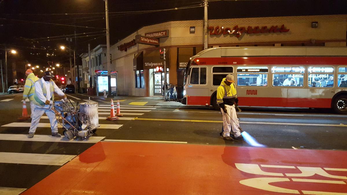Workers in safety vests marking red transit lane on Mission Street at night with passing streetcar.