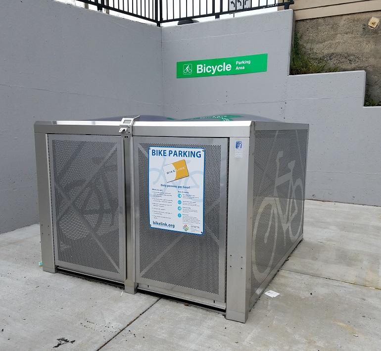 Metal bike lockers with mesh panels in a concrete parking area under a green bicycle parking sign.