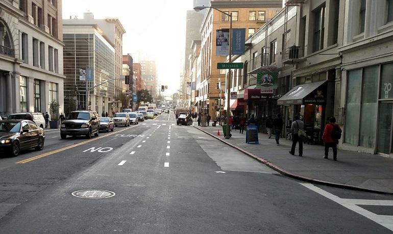 Downtown street with bike lane markings, parked cars, pedestrians, and storefronts along sidewalk.