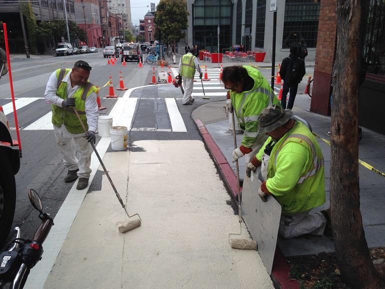 SFMTA workers in safety vests paint a pavement safety zone on a city street with traffic cones and barriers.