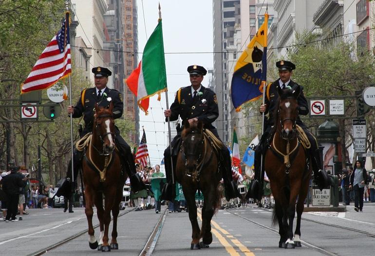 Three mounted police officers on brown horses leading a parade down a San Francisco street with flags and tall buildings.