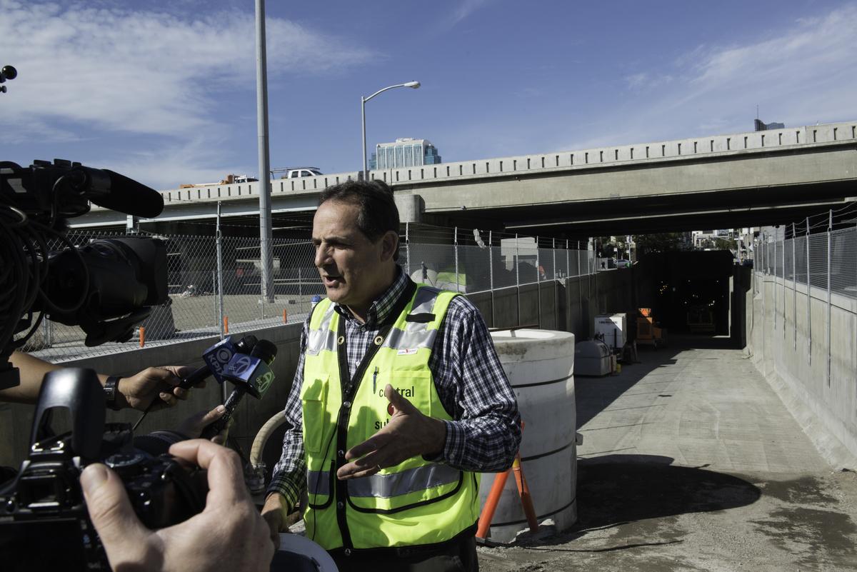 Man in yellow safety vest speaking to media cameras beneath a concrete highway overpass and tunnel portal.