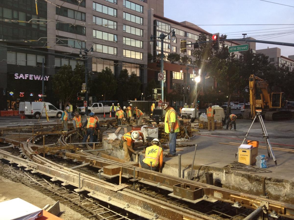 Construction crews in safety vests work on light rail tracks at 4th and King Street at dusk.