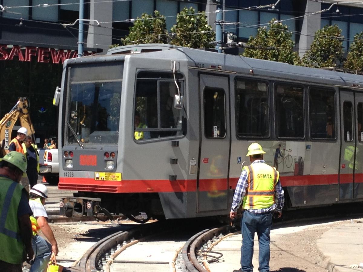 Gray and red Muni light rail train with workers in safety vests and hard hats at 4th and King construction site.