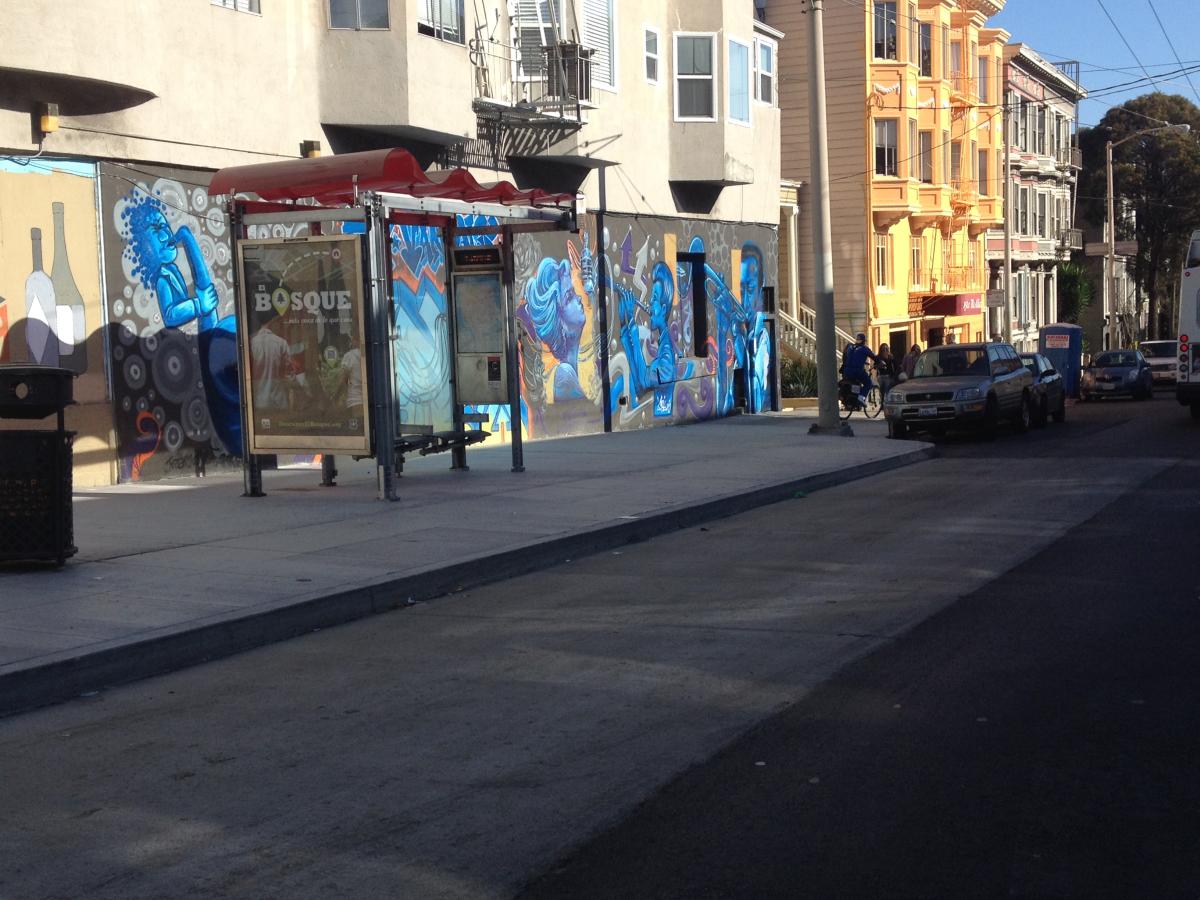 Bus shelter with blue graffiti art on McAllister Street at Divisadero Street, with residential buildings and parked cars visible.