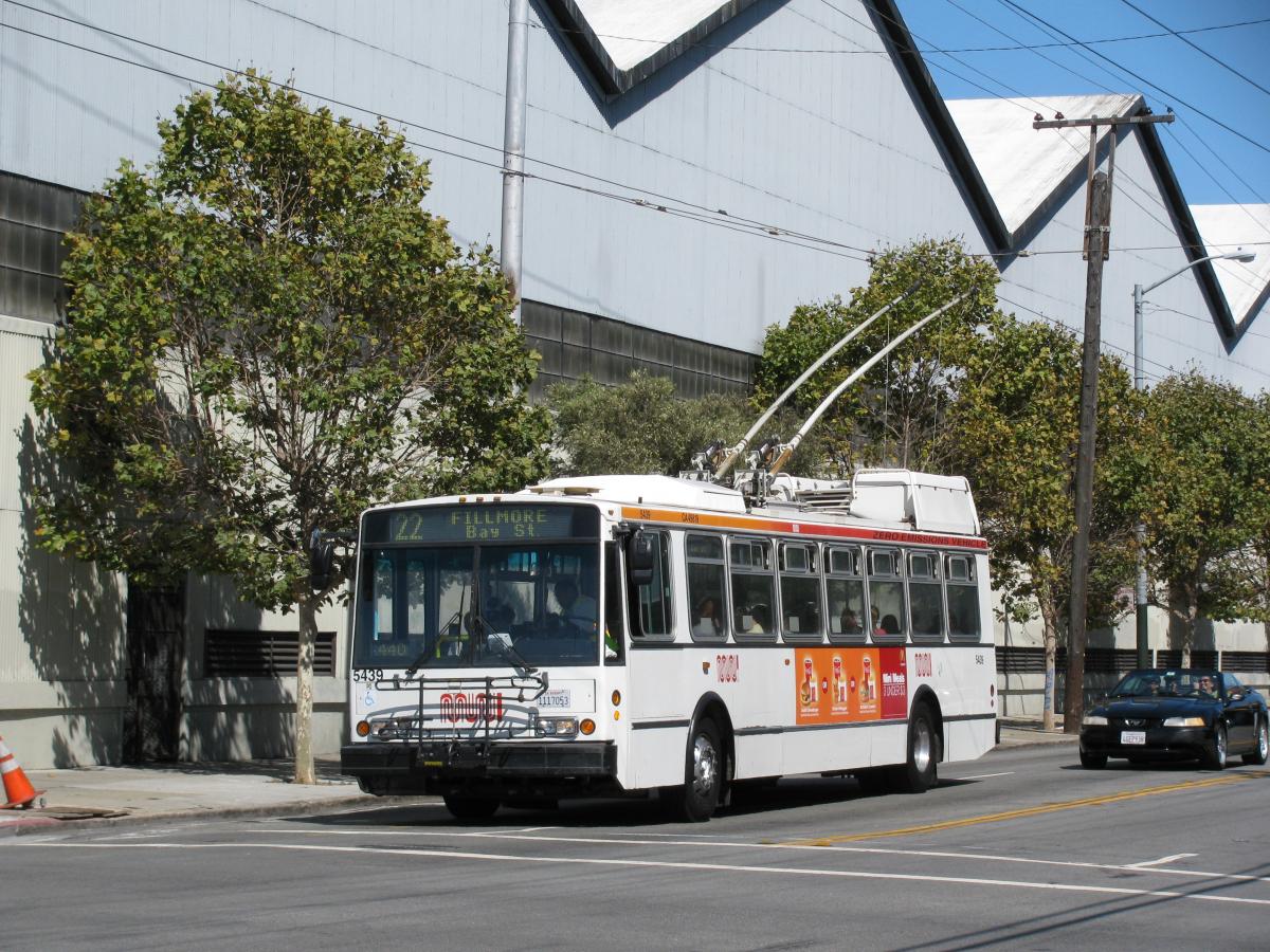 White and orange Muni bus stopped on city street with overhead electric lines and trees visible.