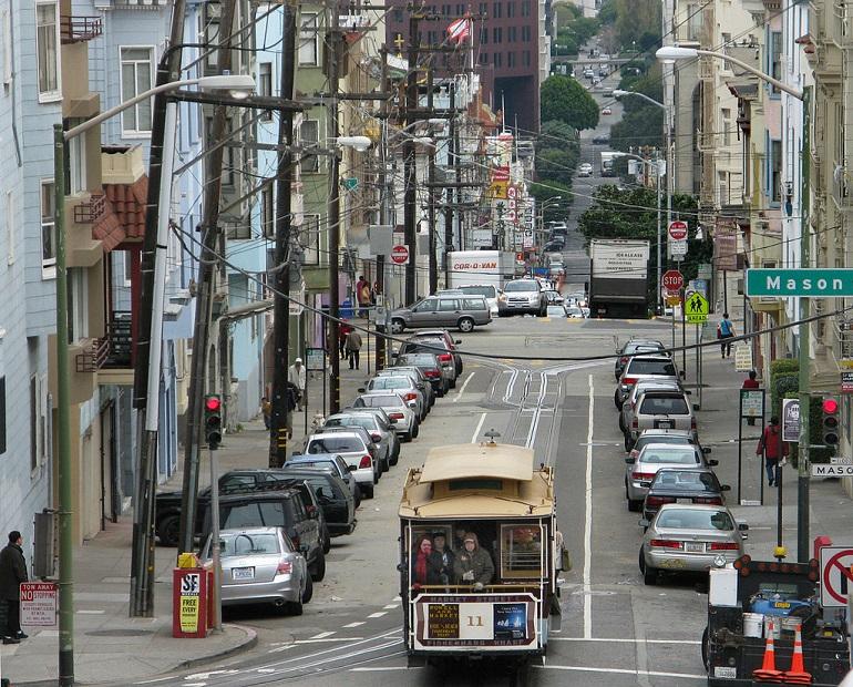 Cable car descending Washington Street at Mason Street in San Francisco, with parked cars lining both sides and traffic in the background.
