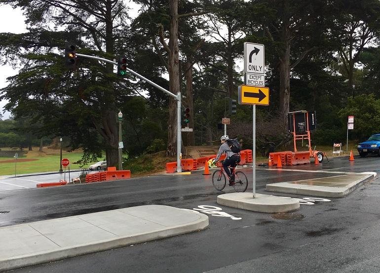 Cyclist riding through opening in traffic island at intersection with orange construction barriers and bicycle signal overhead.