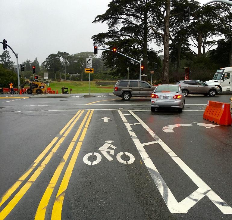 Intersection with bike lane marking, traffic signal, construction equipment and barriers visible on wet pavement.